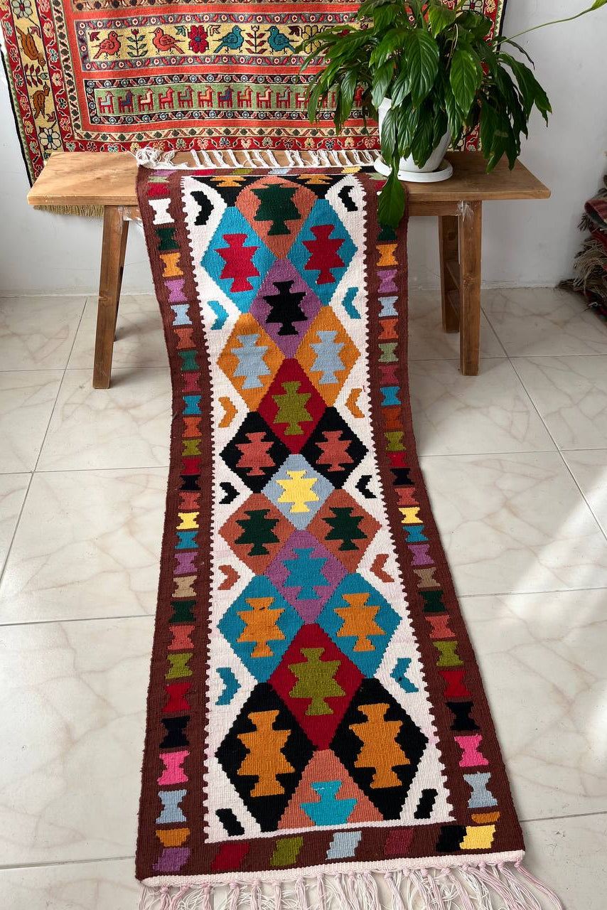 Colorful patterned rug on a wooden table with a plant and tapestry in the background