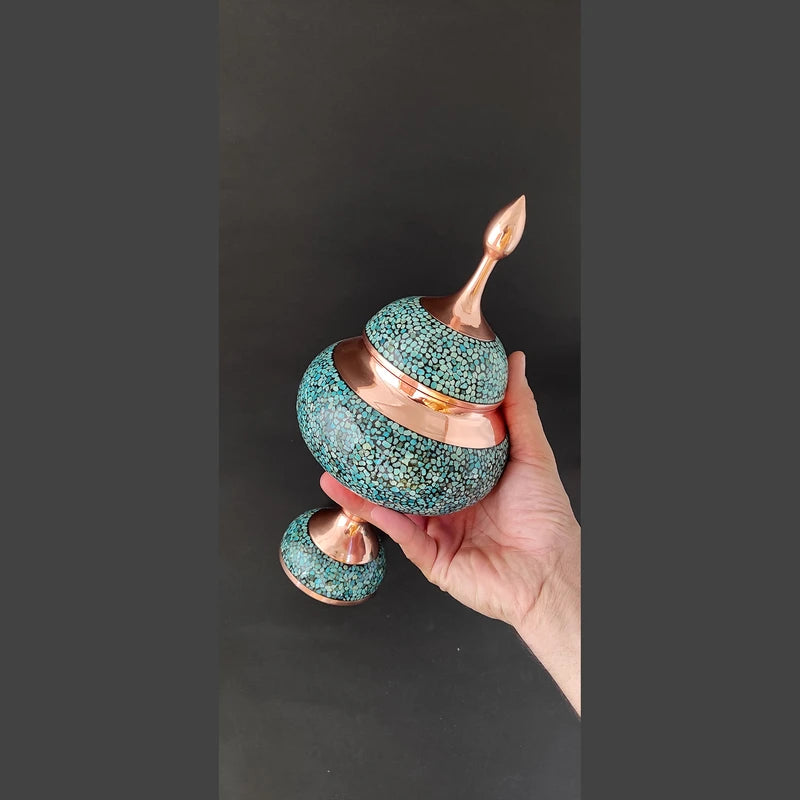 Hand holding a decorative item with turquoise and gold design against a dark background