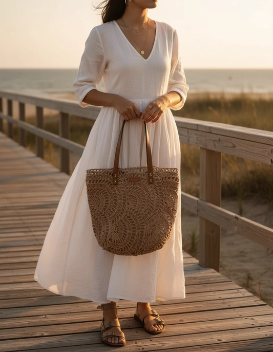 Woman in a white dress holding a brown woven bag on a wooden dock by the water.